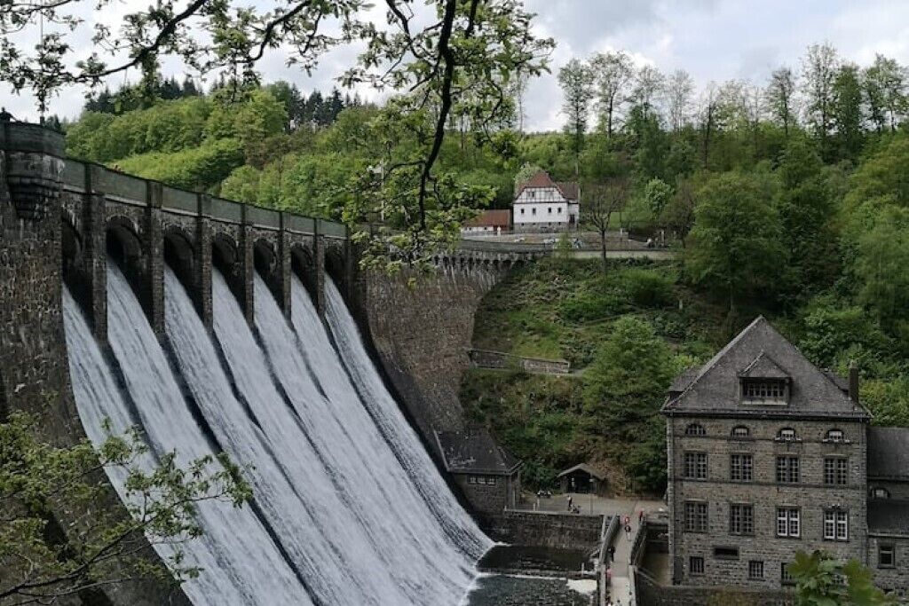 Eine große Staumauer, über die Wasser fließt, umgeben von üppigem Grün und einem historischen Gebäude.