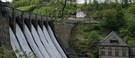 Eine große Staumauer, über die Wasser fließt, umgeben von üppigem Grün und einem historischen Gebäude.