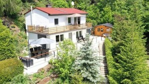 A two-story house with a red roof, a balcony, and a small attached wooden structure.