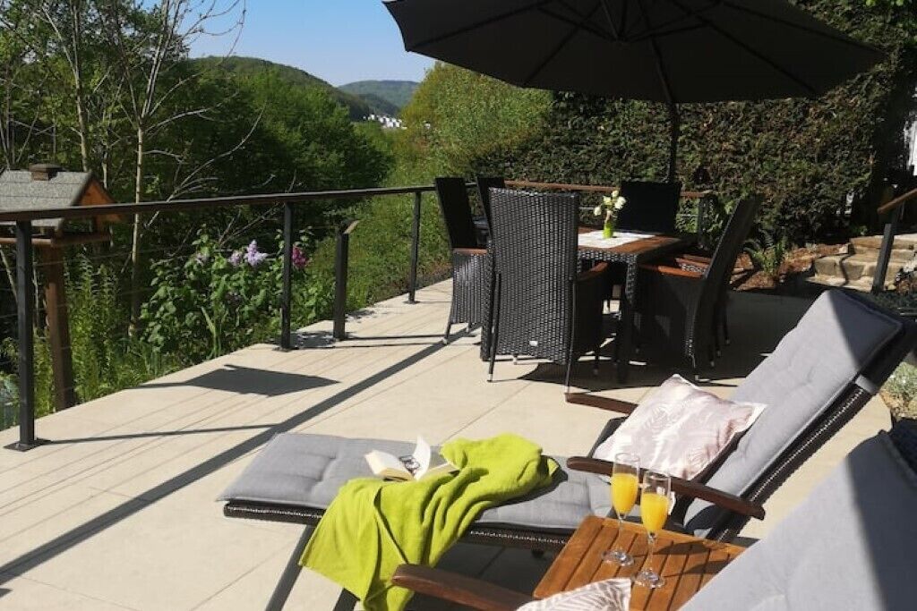 An outdoor seating area with lounge chairs and a table, surrounded by greenery and a view of hills.