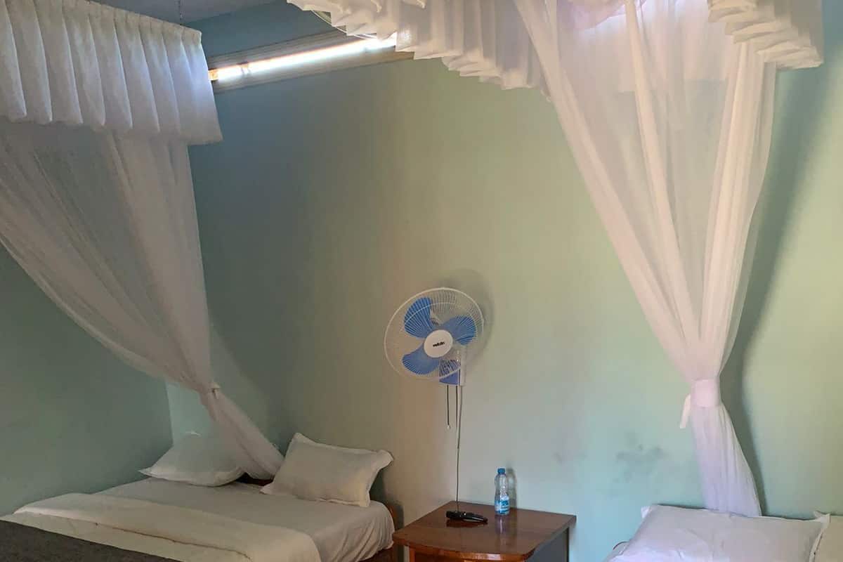A room with two wooden beds, white bedding, a standing fan, and a nightstand with a water bottle.