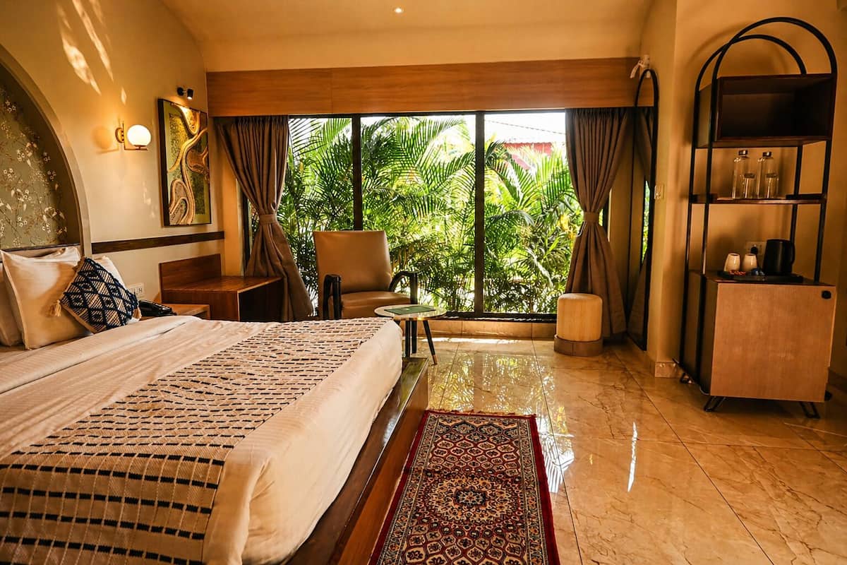 A hotel room with a bed, a desk, a chair, a small table, a sofa, a cabinet, and a view of greenery outside.
