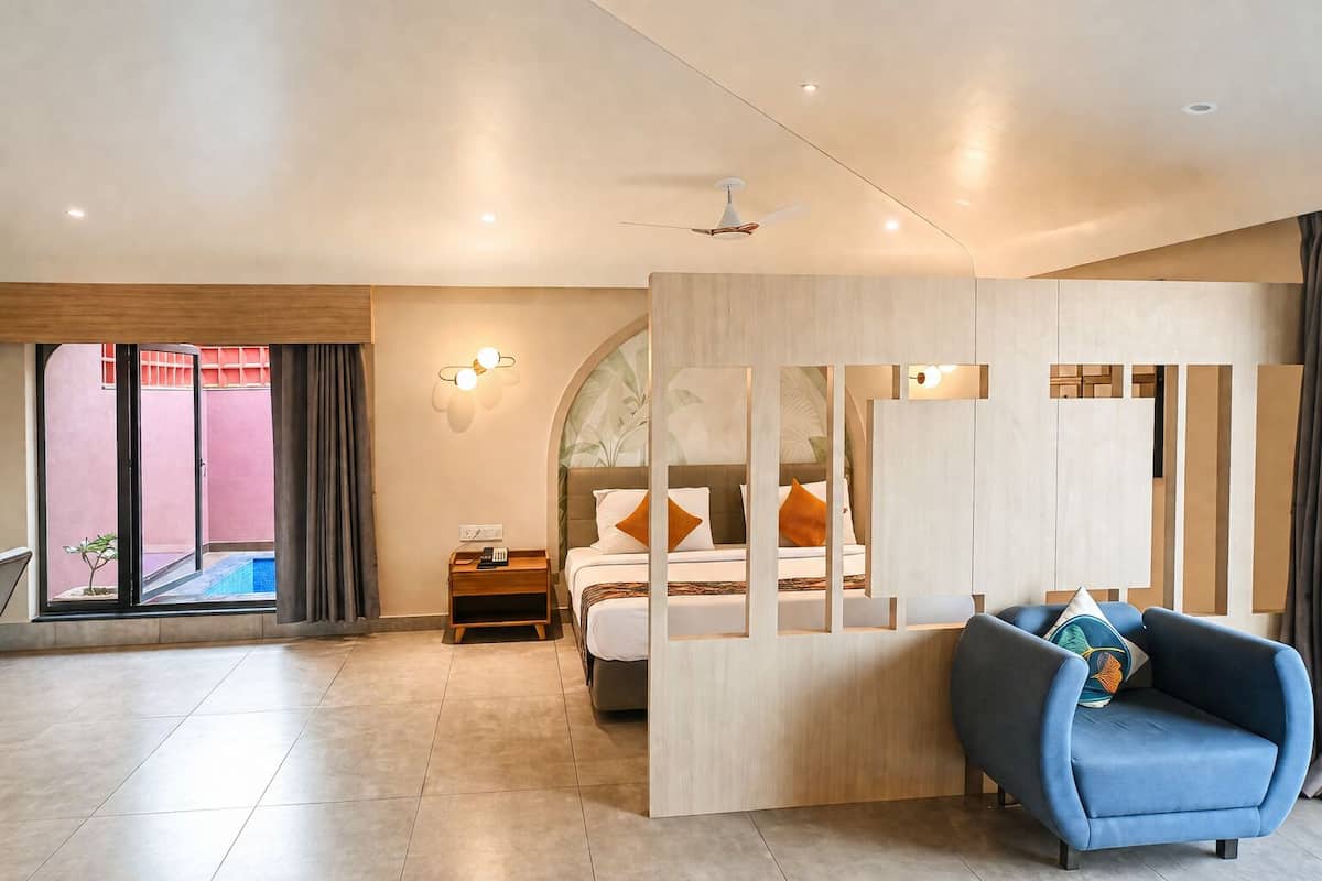 A modern hotel room with a bed, a blue sofa, a wooden partition, a ceiling fan, and a view of an outdoor area.