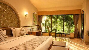 A hotel room with a large bed, a desk, a chair, and a view of greenery outside.