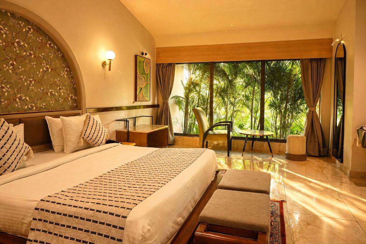 A hotel room with a large bed, a desk, a chair, and a view of greenery outside.