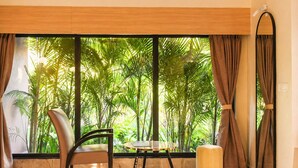A room with a large window, a chair, a small table, and a round ottoman.