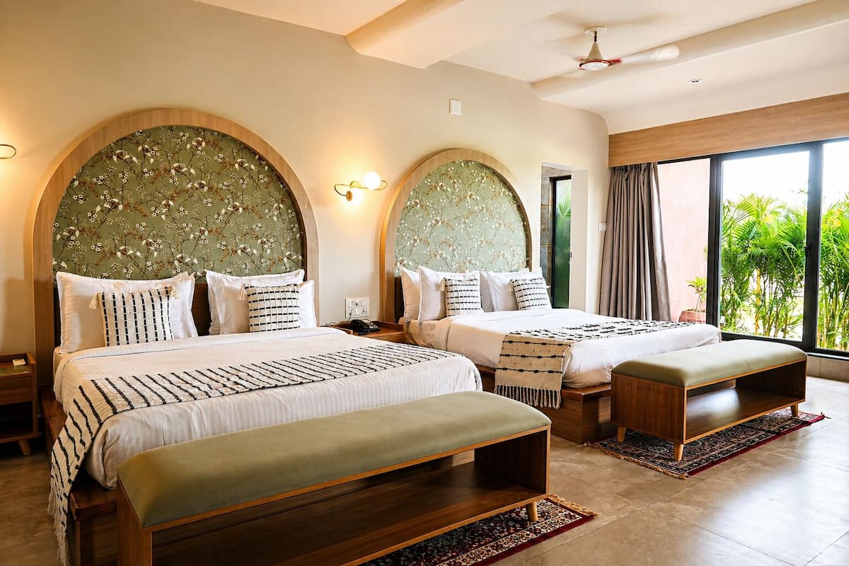 A hotel room with two beds, a bench, a large window with a view of greenery, and a ceiling fan.