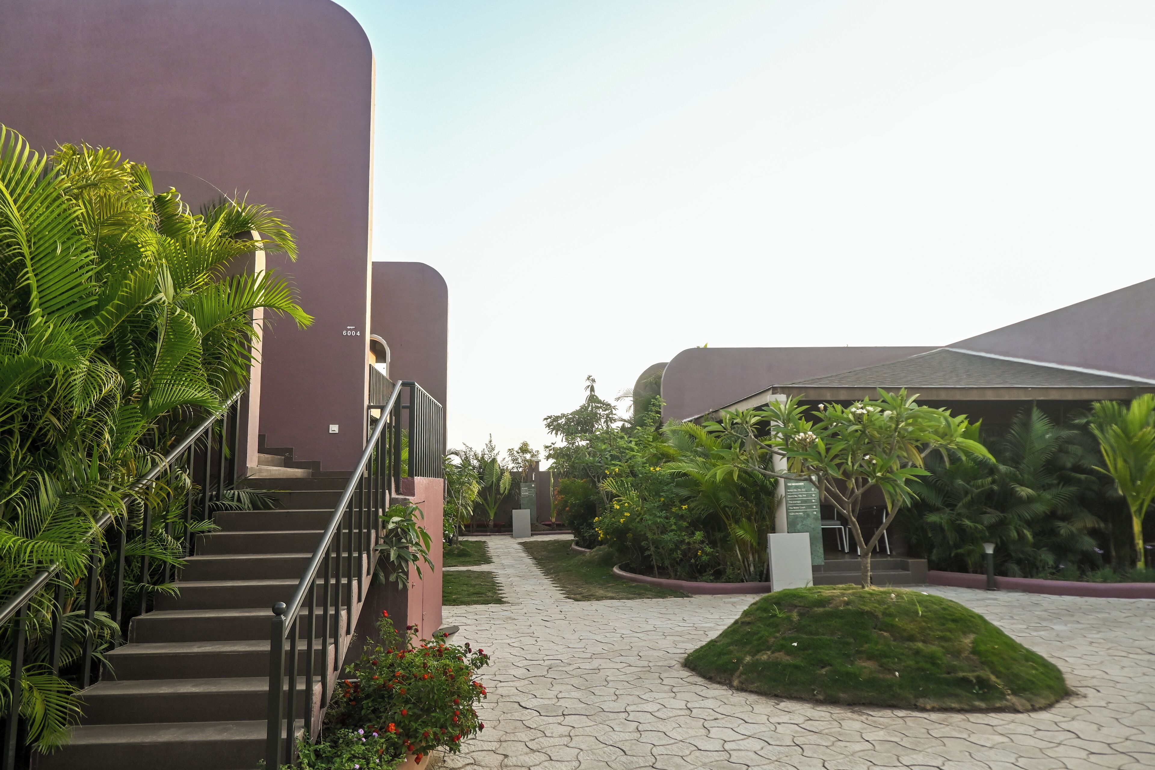 A pathway leading to a building with a staircase, surrounded by greenery and a small hill.