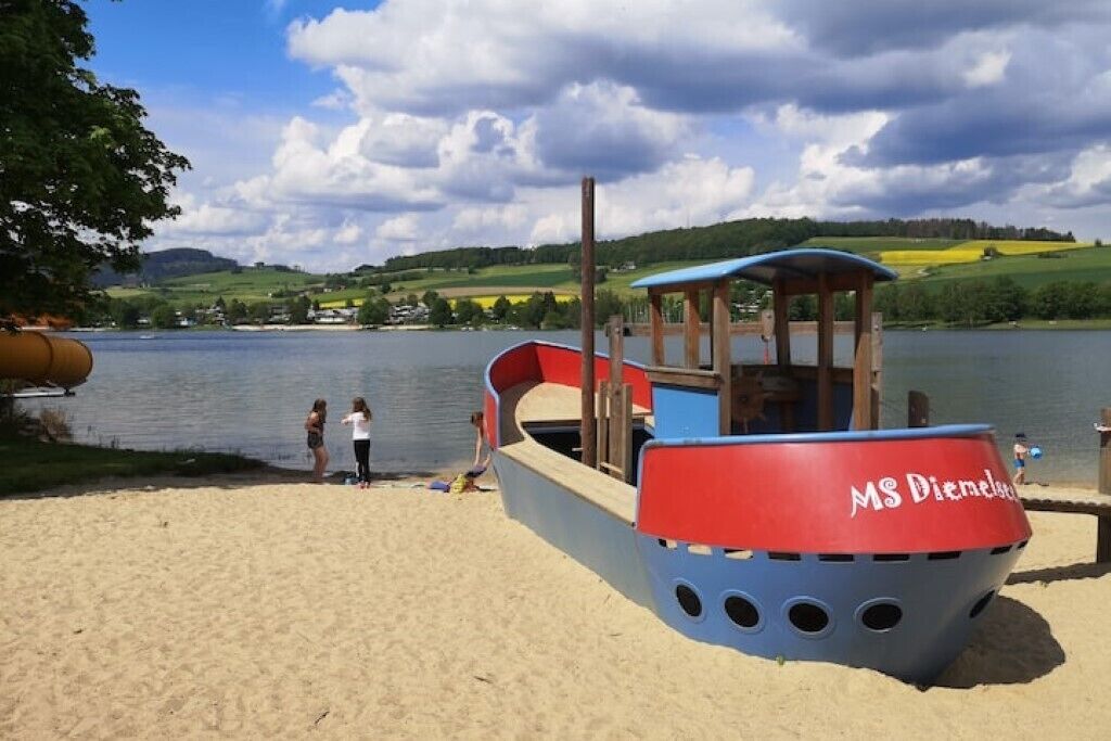 Eine bootförmige Spielanlage mit Kabine und Rutschen, auf einem Sandstrand an einem See gelegen.