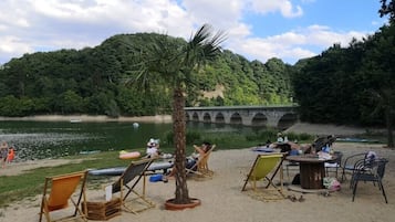 Una playa con sillas de descanso y una palmera, un puente al fondo y una masa de agua tranquila.