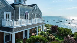 Une grande maison à deux étages, dotée d’un balcon donnant sur une étendue d’eau, et entourée d’une végétation luxuriante.