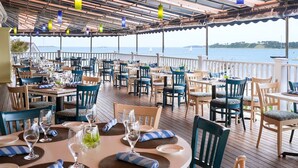 Un espace extérieur abrité, aménagé pour les repas, avec plusieurs tables et chaises, donnant sur une étendue d’eau.