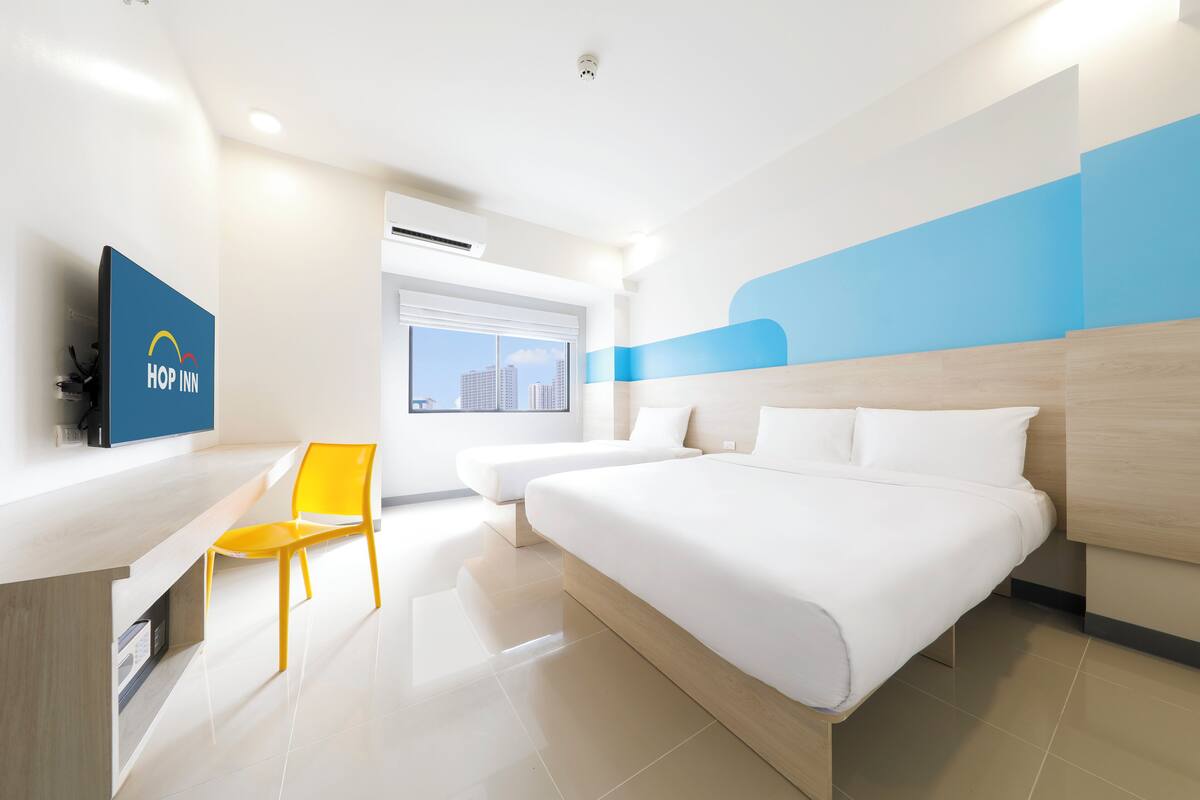 A modern hotel room with a bed, a desk, a chair, and a television displaying the hotel name.
