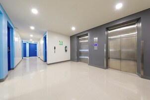 Corredor moderno e bem iluminado, com portas azuis, piso limpo e um elevador.
