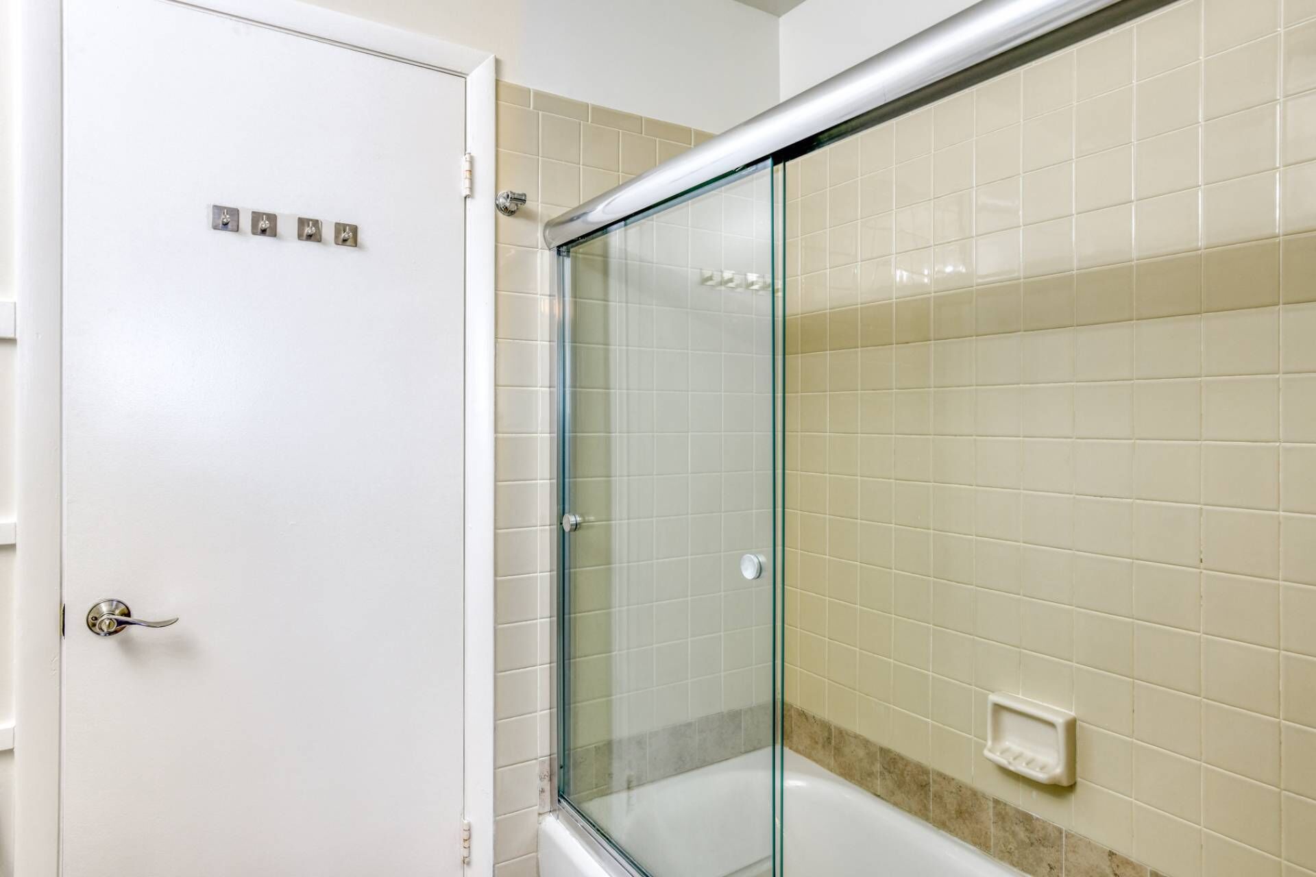 A bathroom with a glass shower enclosure, a white door, and beige tiled walls.