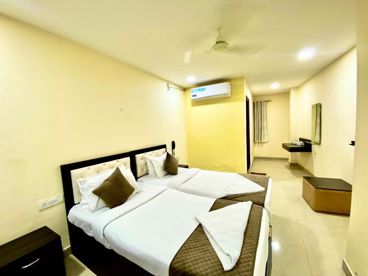 A hotel room with two beds, a small bedside table, a nightstand, a mirror, a sink, and an air conditioning unit.