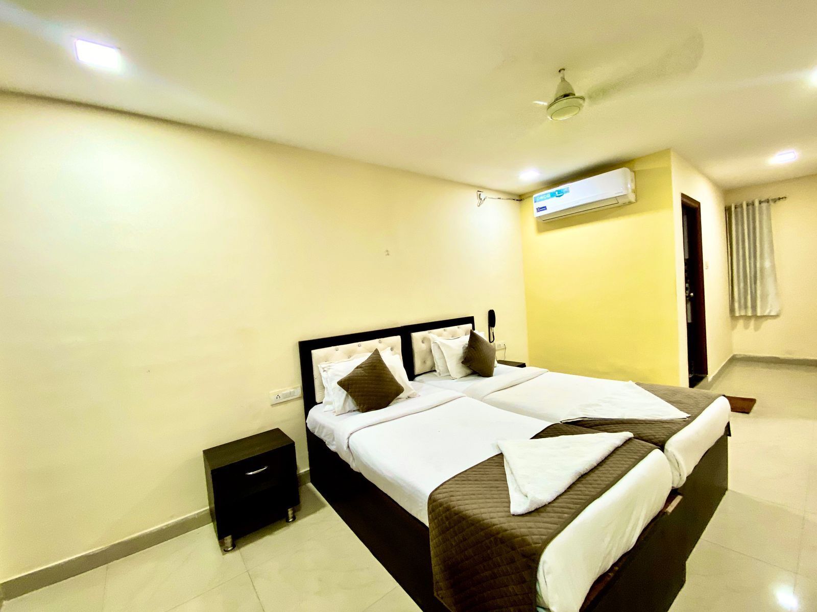 A hotel room with a double bed, a bedside table, an air conditioning unit, and a ceiling fan.