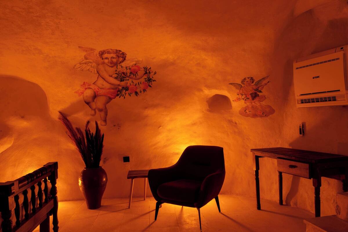 Une pièce décorée d’une fresque représentant des anges, avec une plante en pot, une chaise, une petite table et un tableau blanc.