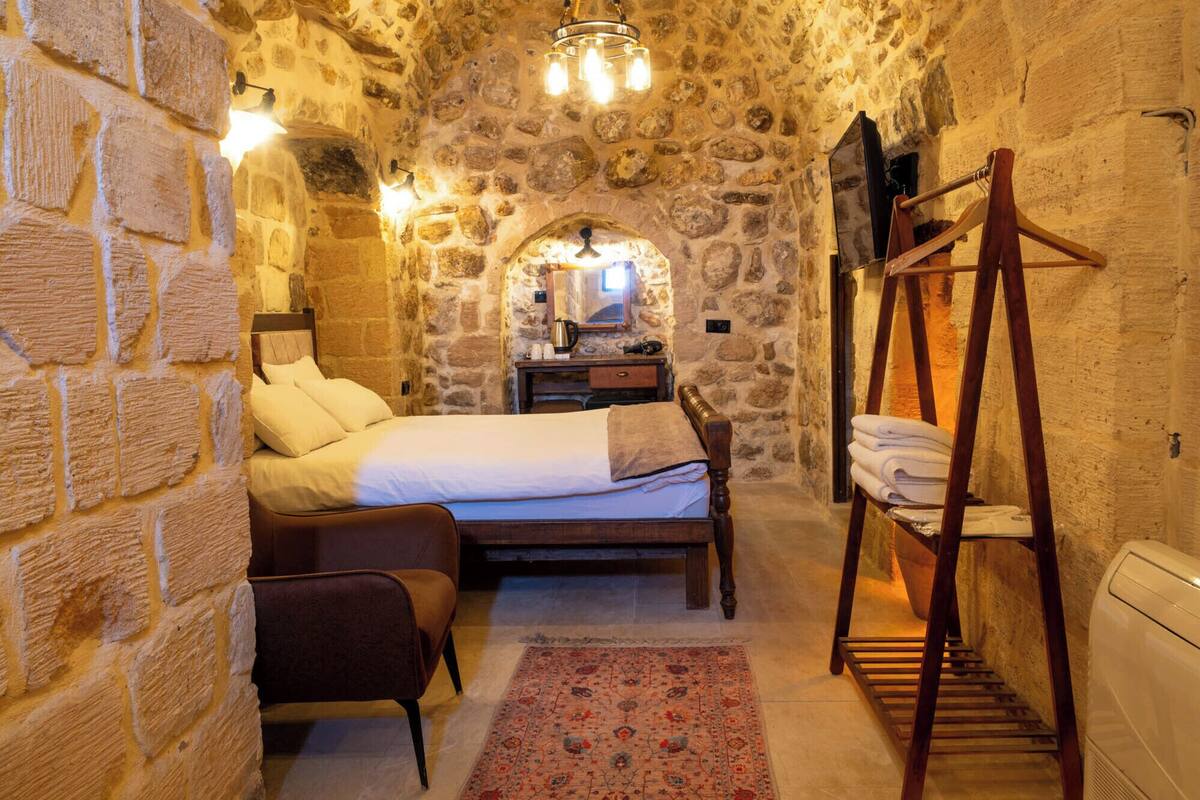 Une pièce aux murs de pierre, avec un lit, une chaise en bois, un étendoir en bois et une petite table sur laquelle se trouve une bouilloire.