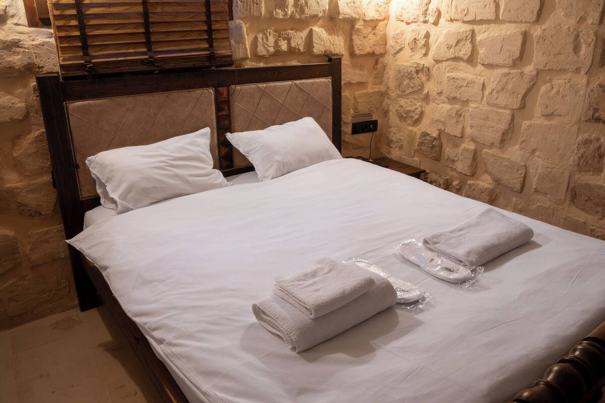 Un lit bien fait, avec du linge de lit blanc et deux serviettes pliées, adossé à un mur de pierre et doté d’une tête de lit en bois.