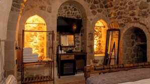 A stone-walled room with a bed, a wooden dresser, a mirror, and a fireplace. - Marvel Otel (Mardin)
