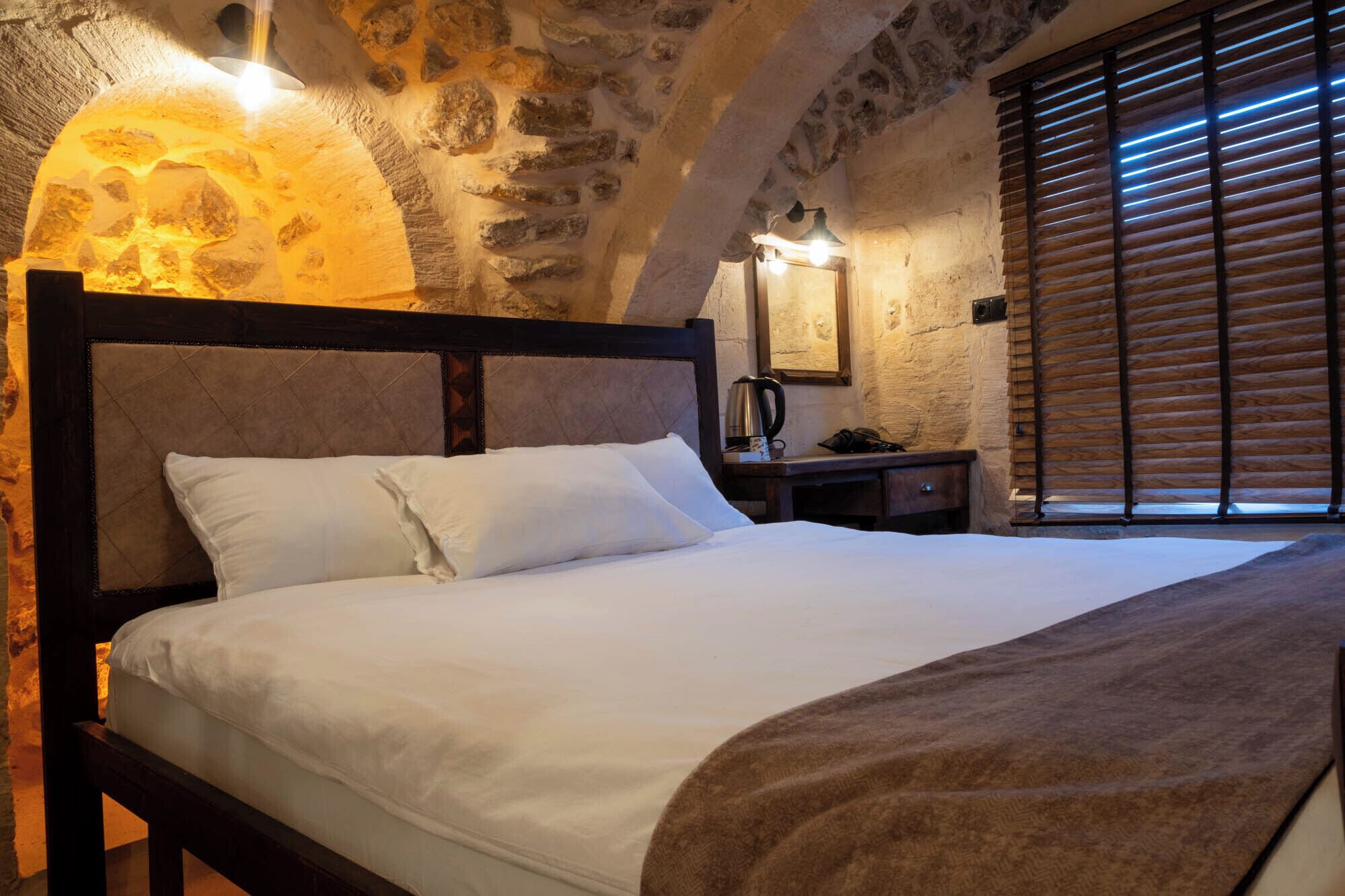 Un dormitorio con pared de piedra, cabecera de madera, cama con ropa de cama blanca, mesita de noche con hervidor y ventana con persianas de madera.