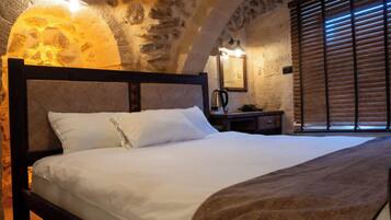Un dormitorio con pared de piedra, cabecera de madera, cama con ropa de cama blanca, mesita de noche con hervidor y ventana con persianas de madera.
