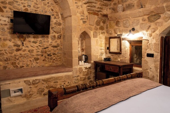A stone-walled room with a bed, a flat-screen TV mounted on the wall, a wooden bench, a small table with a mirror, and a wall-mounted light fixture.
