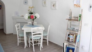 Un comedor con una mesa y sillas blancas, un jarrón con flores y una estantería con libros y objetos decorativos.
