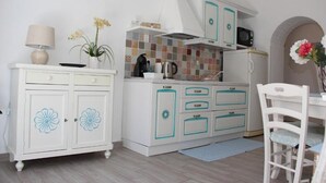 Cocina compacta con alacenas blancas, un microondas y una tetera. Hay un pequeño comedor con una silla y una mesa.