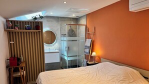 A modern bedroom with a large bed, a glass-enclosed shower, a wooden vanity, and a skylight.
