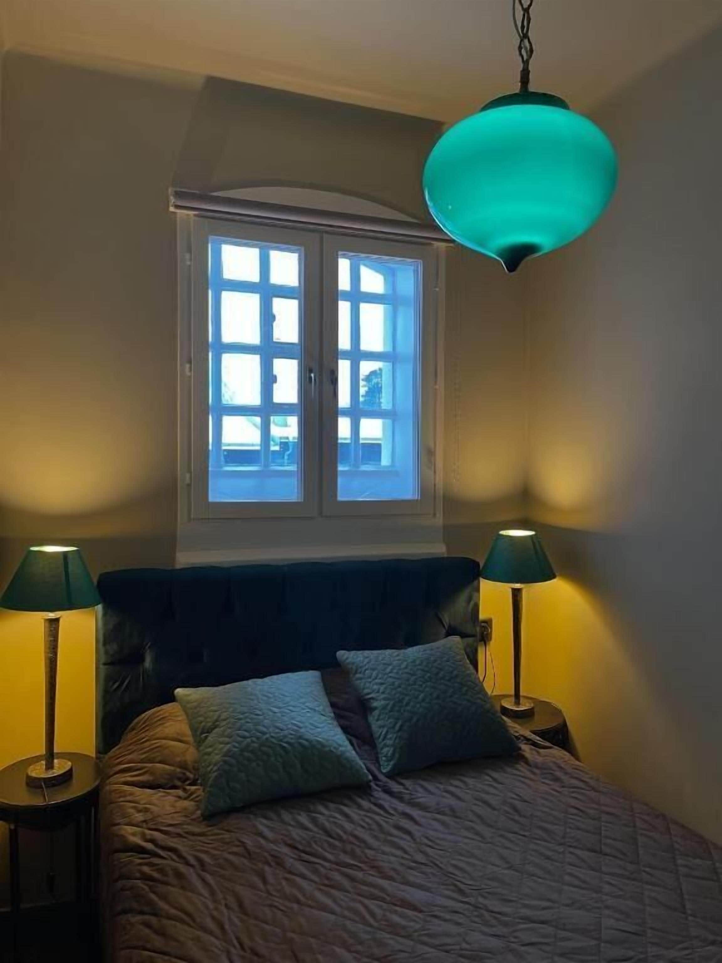 Un dormitorio con una cama, dos lámparas de noche, un ventanal y un candelabro.