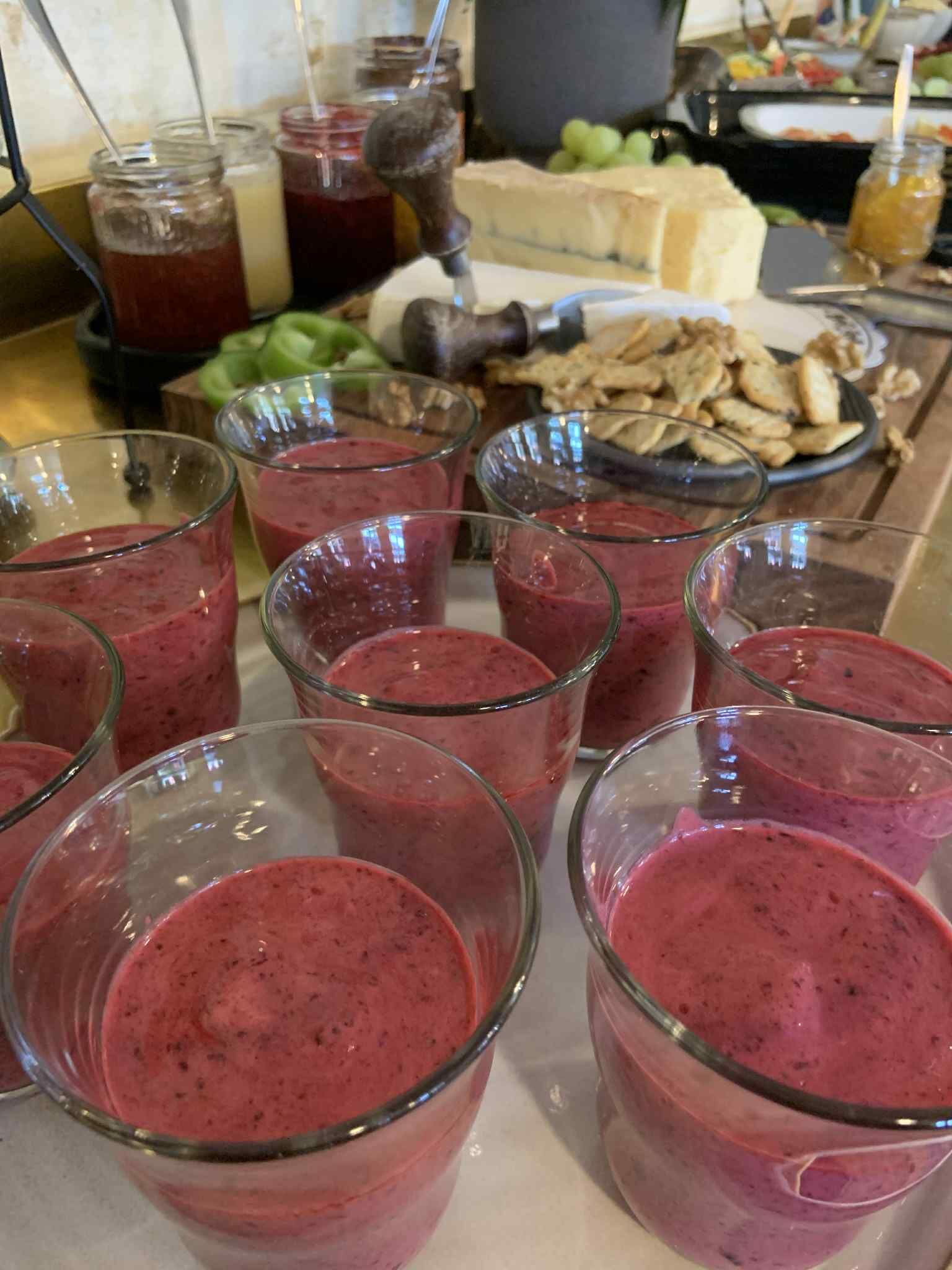 A table with several glasses of pink smoothie and various food items in the background.