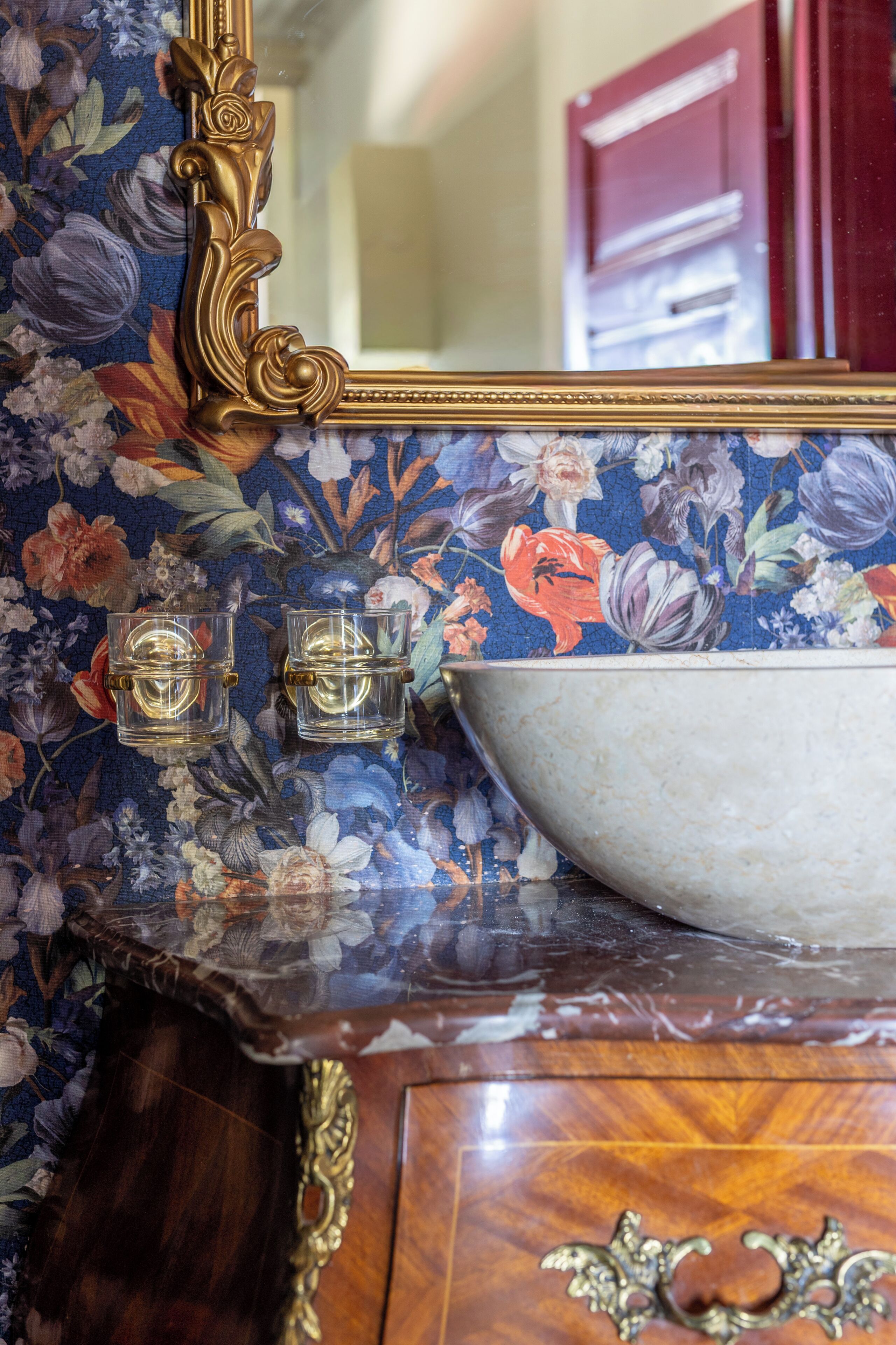 A bathroom with a floral-patterned wall, a marble sink, and a gold-framed mirror.
