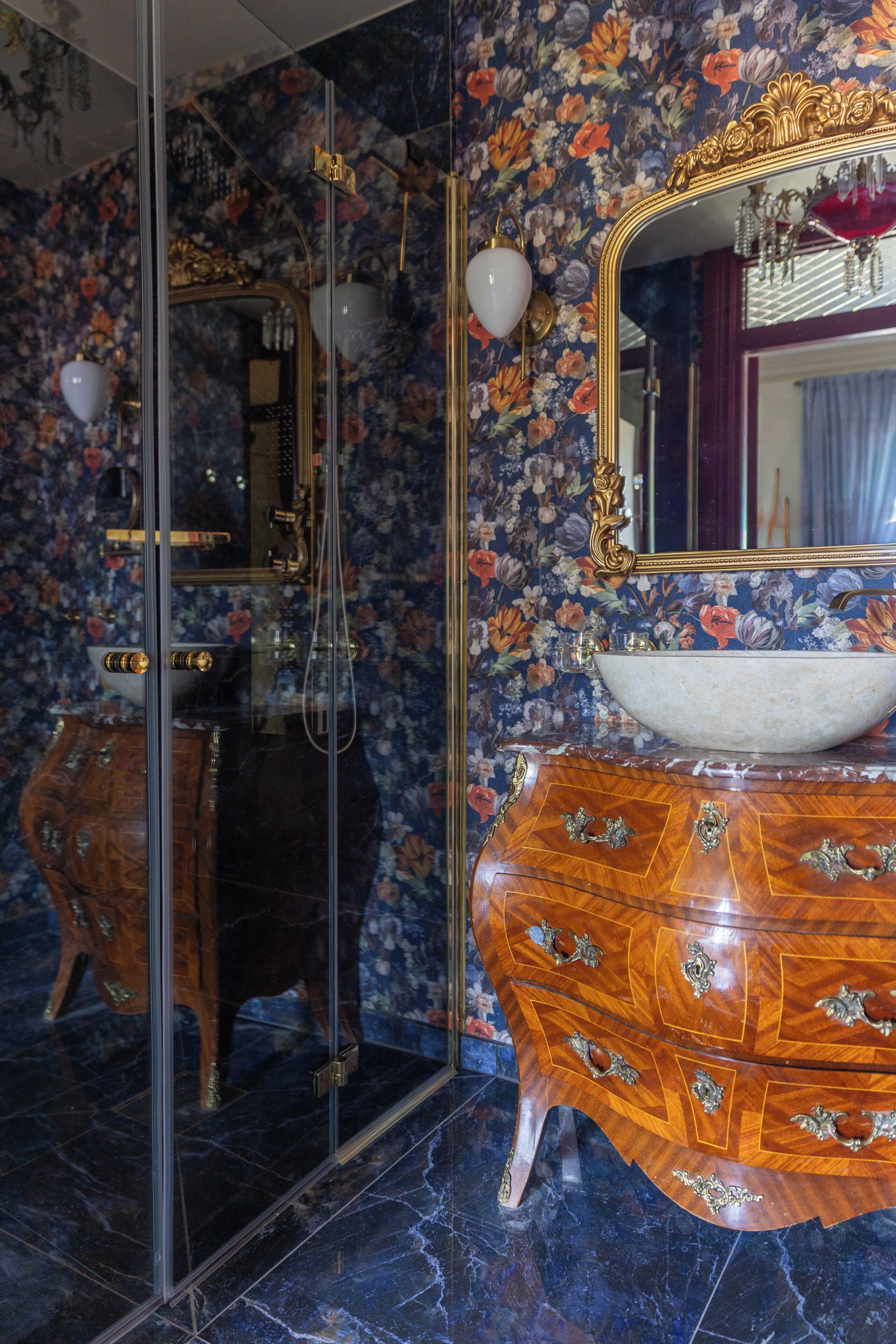 Un baño con piso de mármol, tocador de madera con elegantes pomos, un espejo grande con marco dorado y papel tapiz floral.