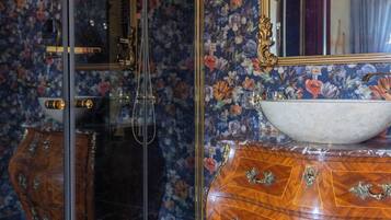 Un baño con piso de mármol, tocador de madera con elegantes pomos, un espejo grande con marco dorado y papel tapiz floral.