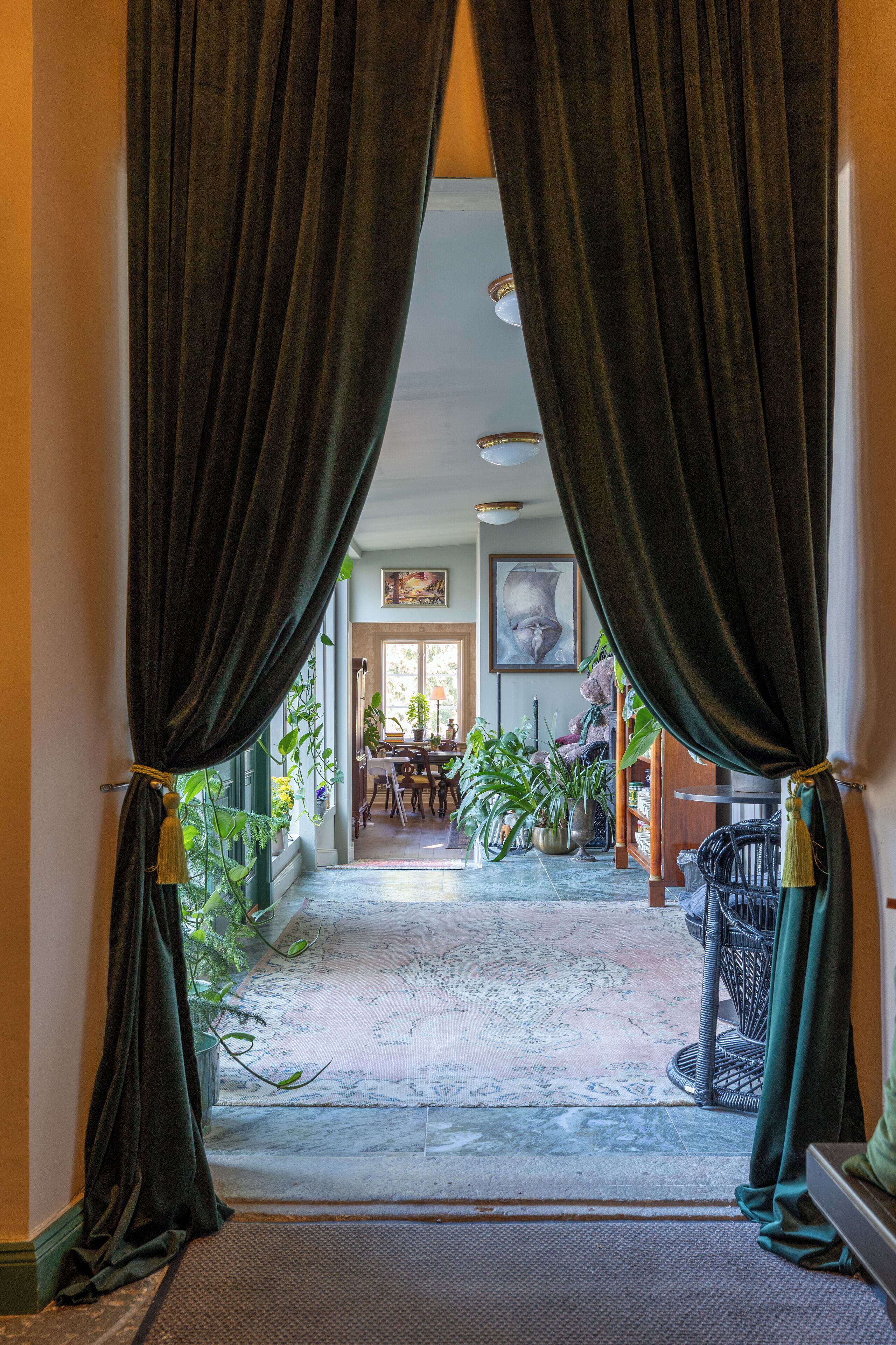 A hallway with dark curtains, a patterned rug, and a dining area with chairs and plants.
