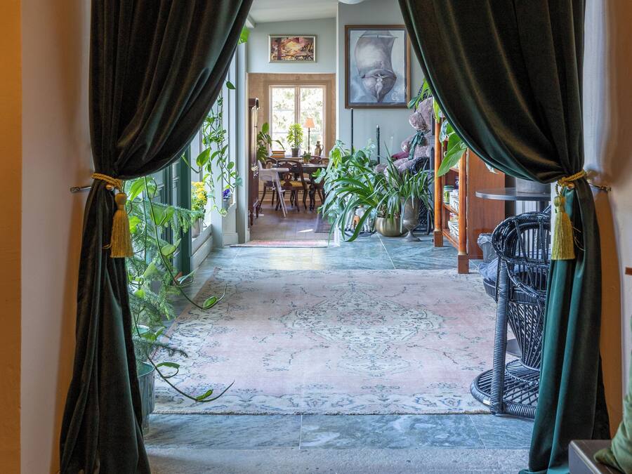 A hallway with dark curtains, a patterned rug, and a dining area with chairs and plants.