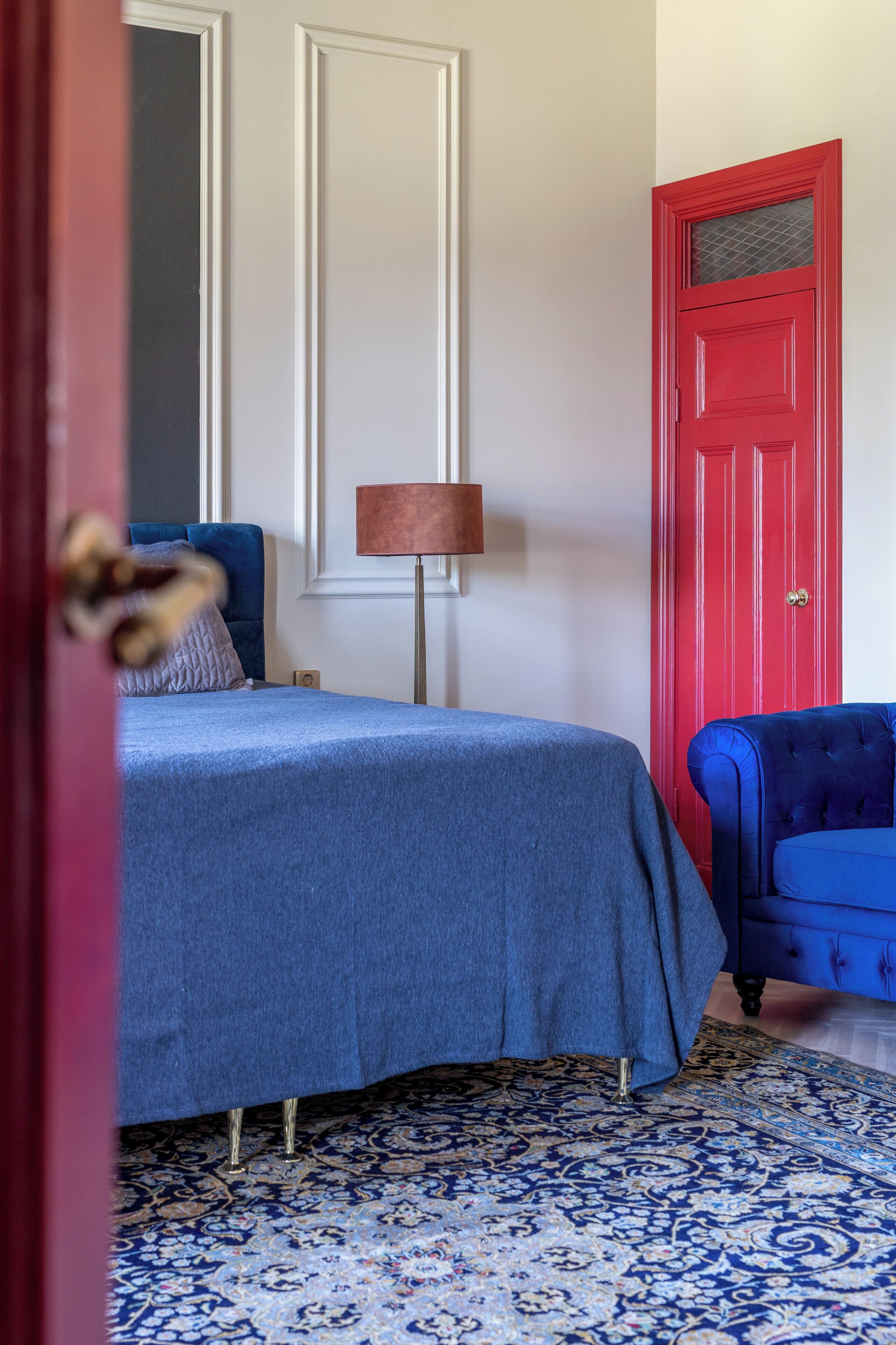 A bedroom with a bed, a blue chair, a red door, and a patterned rug.