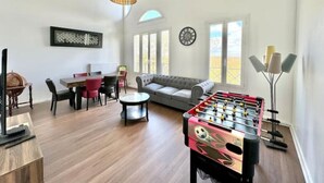 Una sala de estar con sofá, una mesa de comedor con sillas, una mesa de fútbol de mesa y un amplio ventanal con vistas.