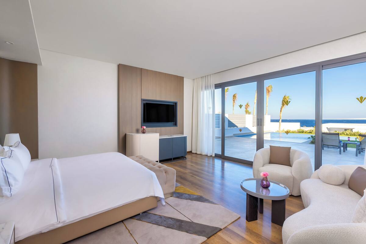 A hotel room with a bed, a TV, a desk, a chair, a coffee table, and a balcony with a view of the ocean.