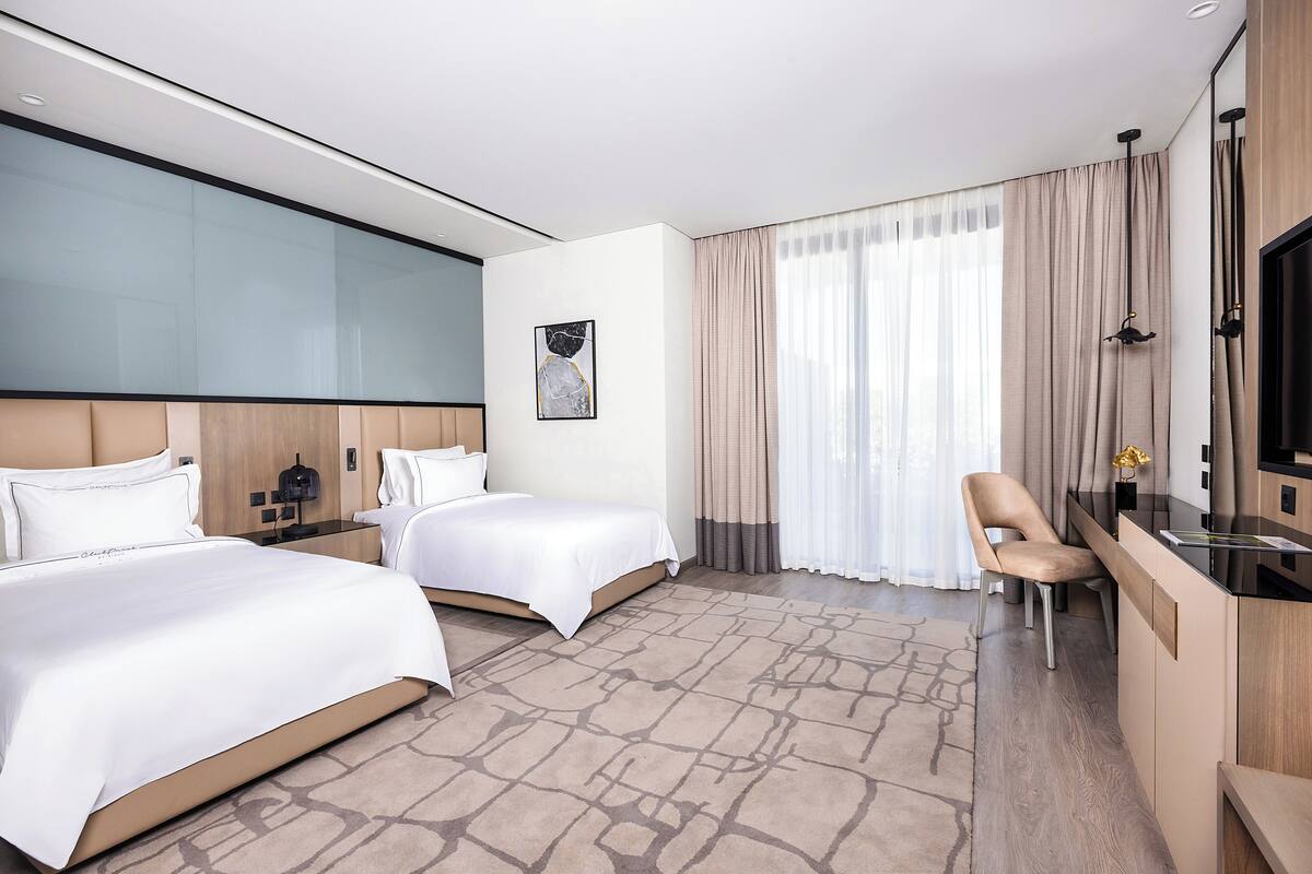 A hotel room with two beds, a desk, a chair, a TV, and a large window with curtains.