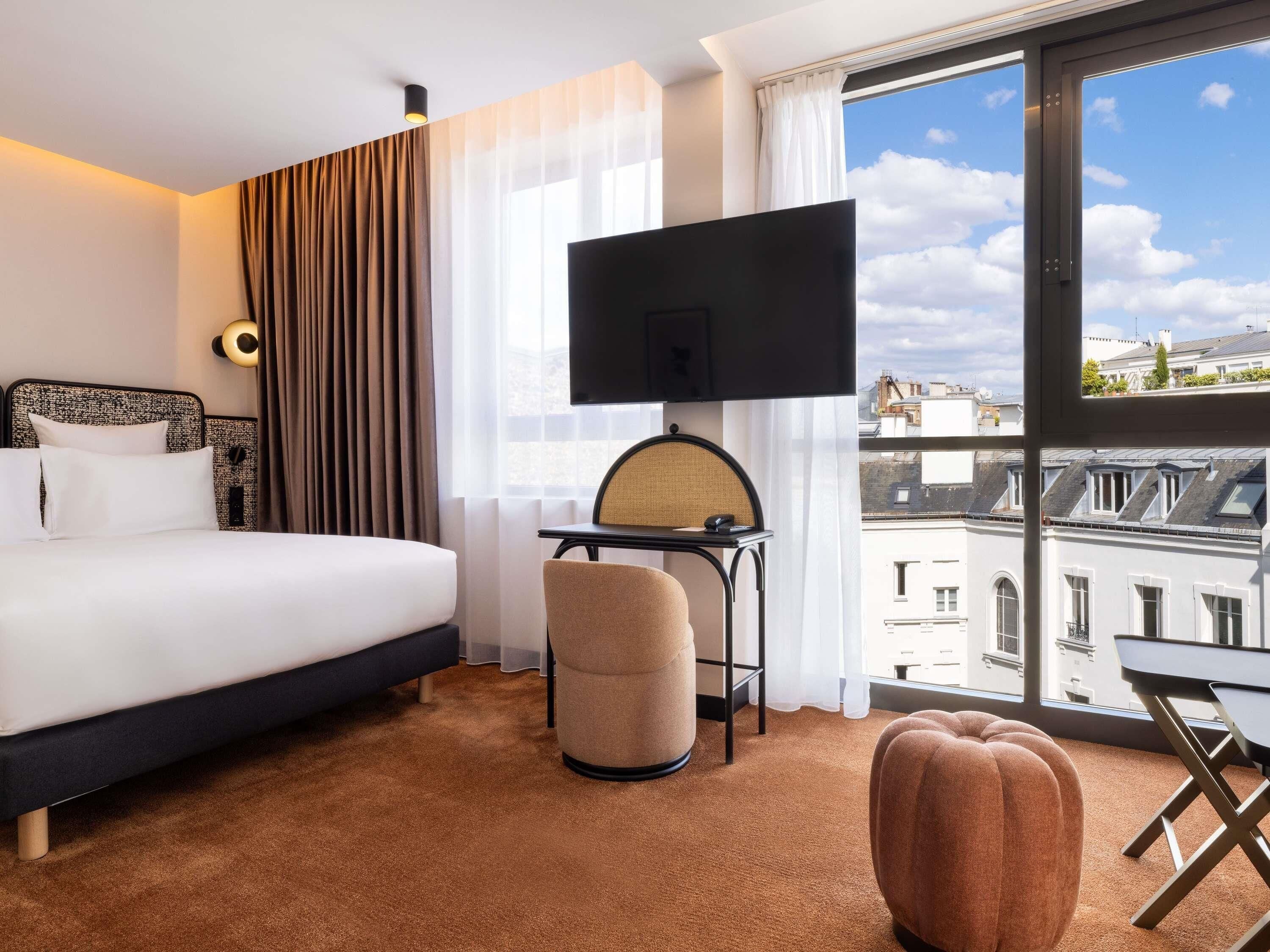 Une chambre d’hôtel comprenant un lit, un bureau avec une chaise, une télévision et une grande fenêtre donnant sur des immeubles.