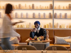 Un restaurant où une personne est assise à une table devant un verre d’eau, un panier de pain et une assiette de nourriture.