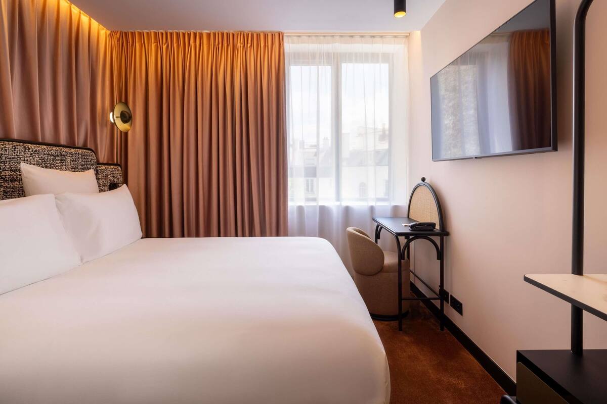 Ett hotellrum med en stor säng, ett skrivbord med en stol, en tv och ett fönster med gardiner.