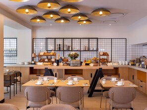 A modern dining area with multiple tables and chairs, a buffet counter with various food items, and pendant lighting.