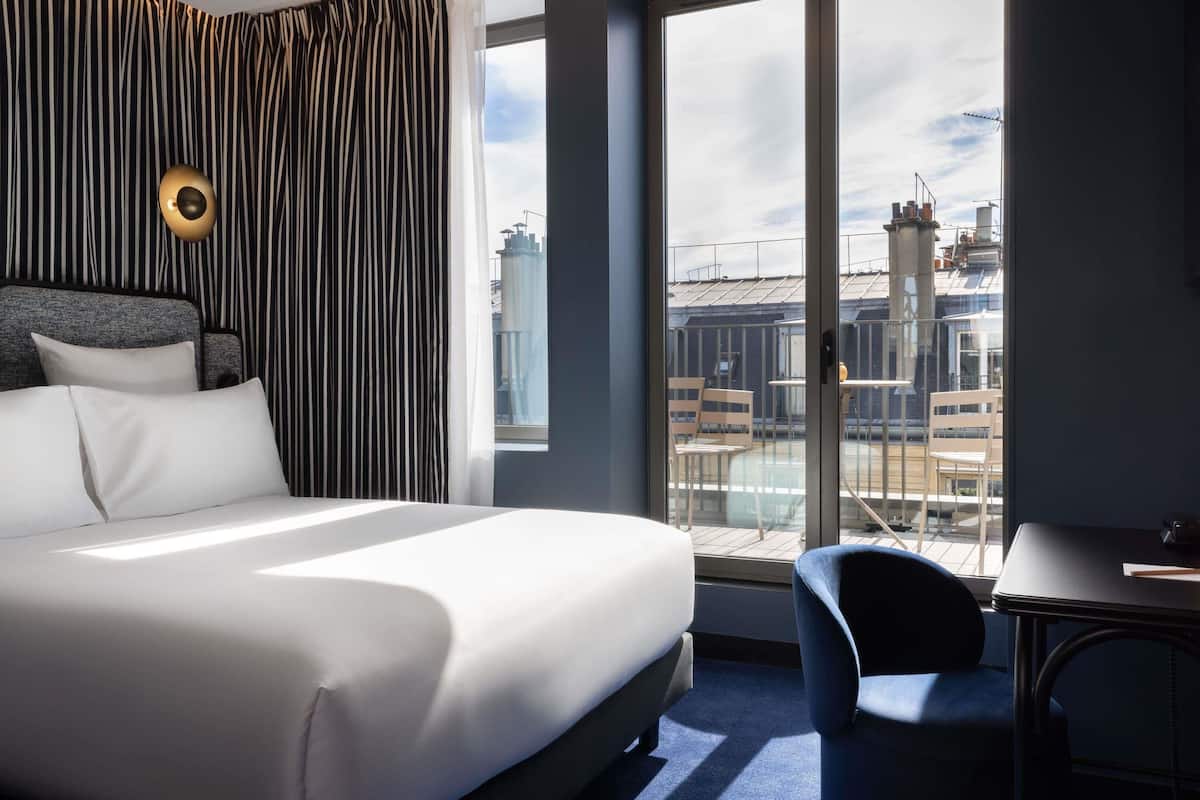 Ett hotellrum med en stor säng, ett skrivbord och utsikt över stadsbilden genom stora fönster.
