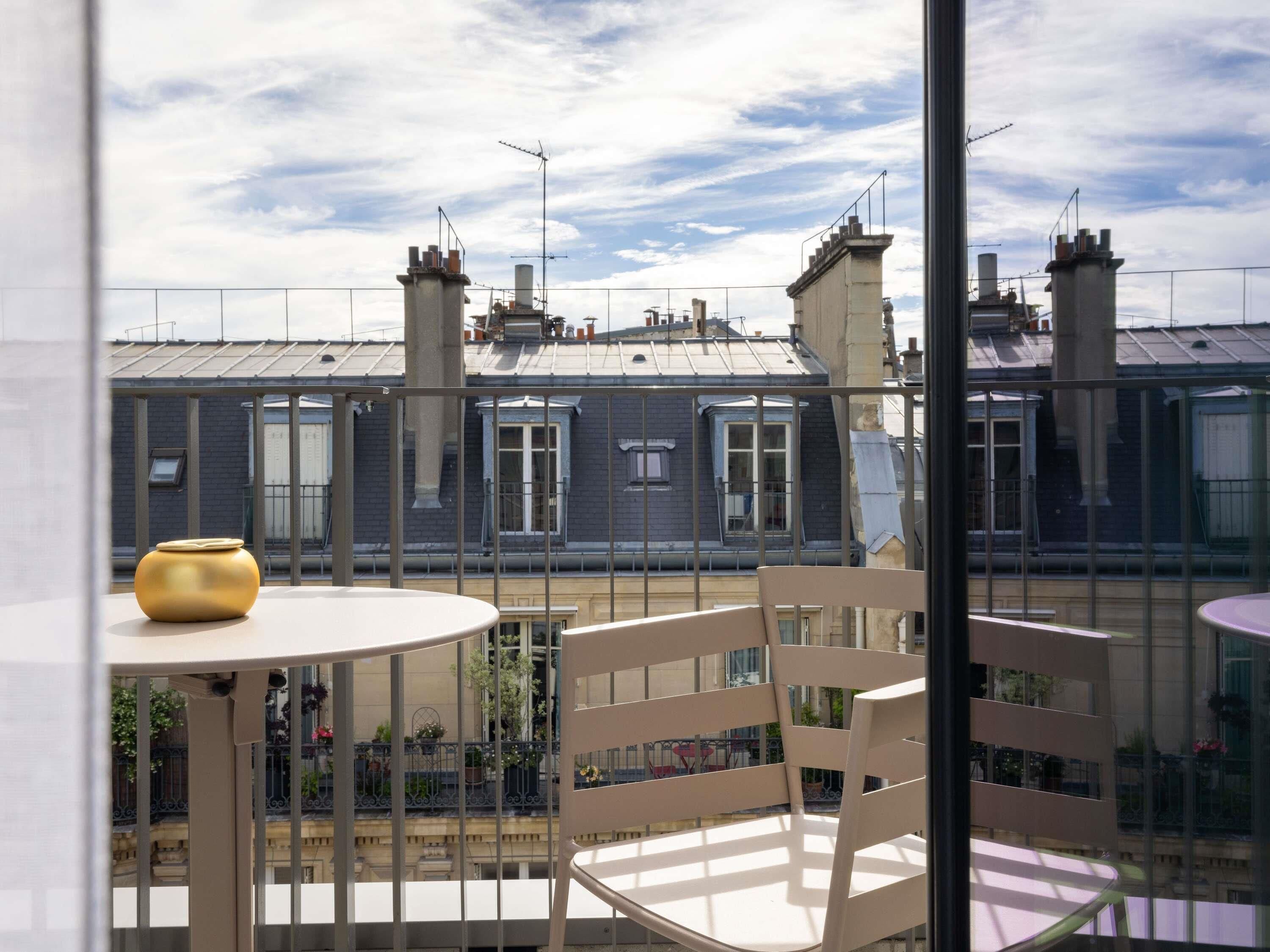 A balcony with a table and chair, a yellow pot, and a view of buildings with chimneys.
