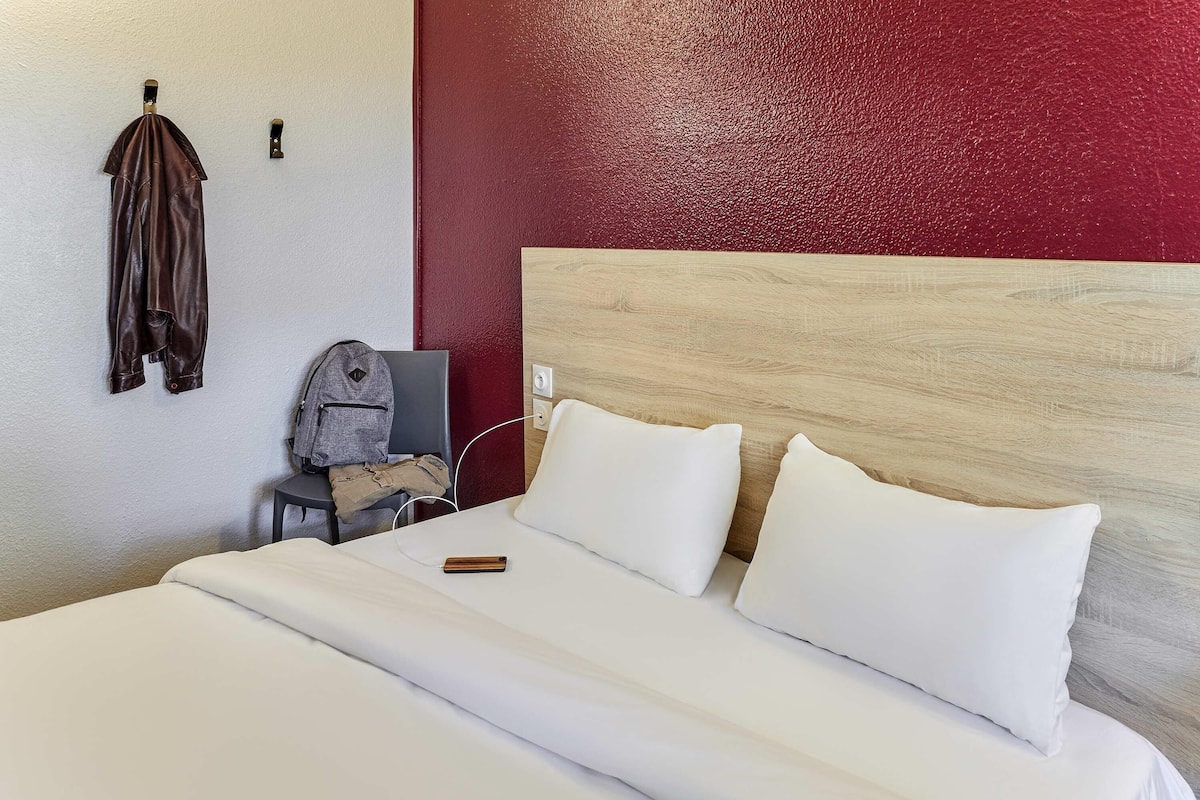 A hotel room with a bed, a chair, a coat rack, and a red accent wall.