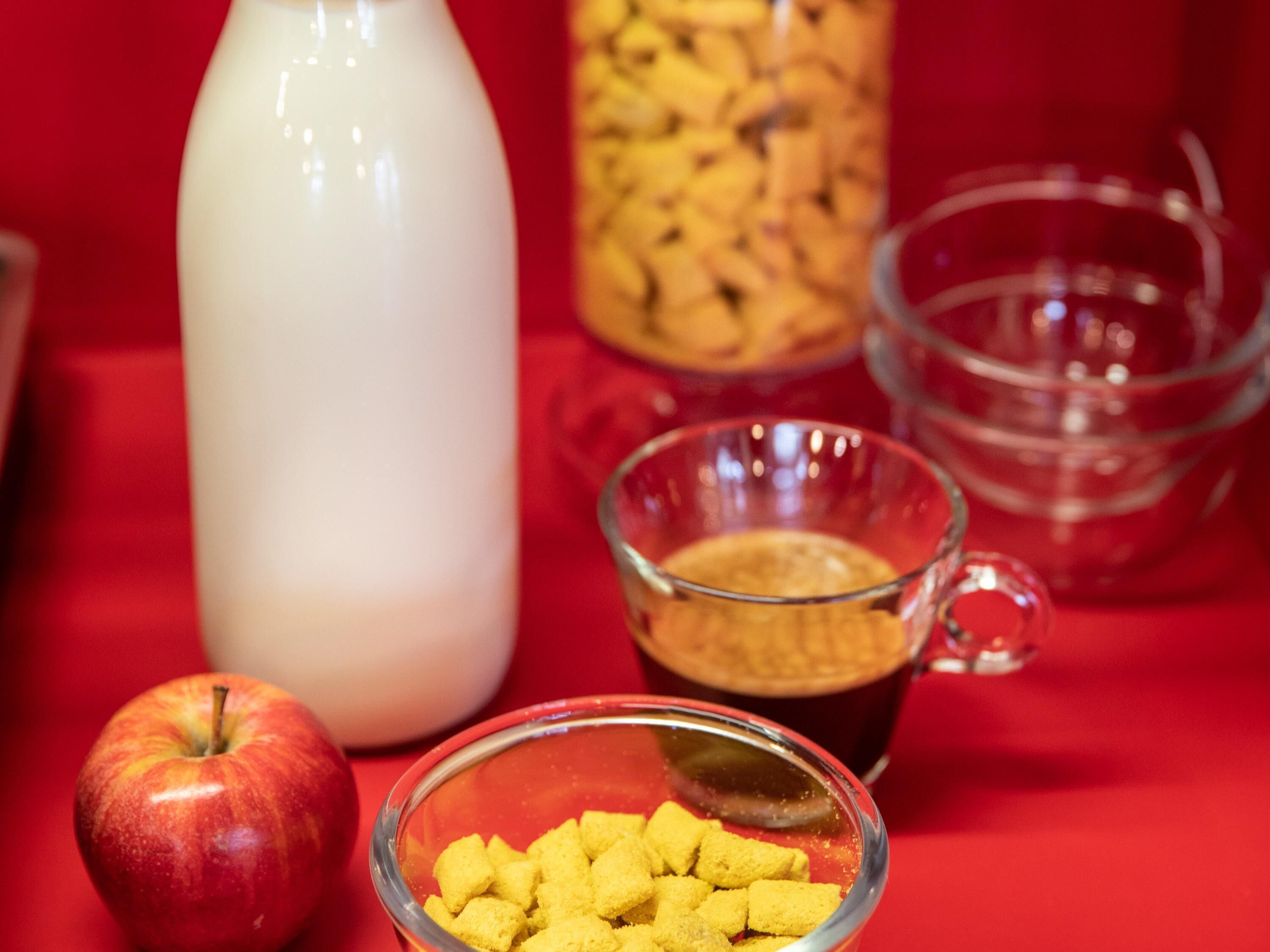 Ein Tisch mit rotem Hintergrund, eine Schale aus Glas gefüllt mit gelben Würfeln, eine Tasse aus Glas mit dunkler Flüssigkeit, eine Karaffe aus Glas und ein roter Apfel.